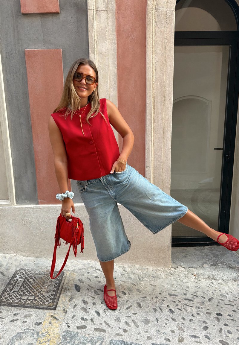 Femme portant un haut rouge sans manches, un pantacourt en denim, des chaussures rouges et des lunettes de soleil, se tient sur une jambe en tenant un sac à main rouge contre un mur texturé.