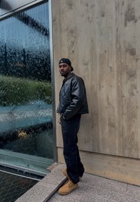 Černá kožená bunda, tmavé džíny a hnědé pracovní boty. Stojí opřený o texturovanou zeď vedle skleněného panelu pokrytého kapkami deště.