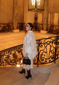 Hvid blondeponcho med detaljerede mønstre, kombineret med cremefarvede bukser og sorte stiletter, holder en lille sort håndtaske, i en udsmykket trappeopgang.