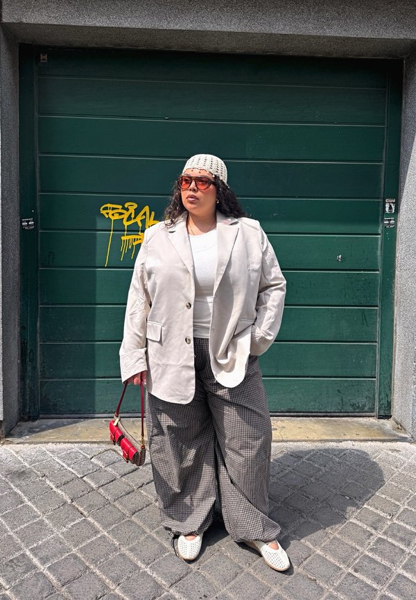 Persona con blazer blanco, pantalones a cuadros, zapatos blancos y un sombrero blanco de crochet, de pie frente a una puerta de garaje verde con grafiti amarillo.