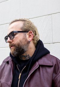 Oseba z brado in kodrastimi blond lasmi, črnimi sončnimi očali, ušesnimi uhani, tetovažo na vratu, oblečena v črno kapuco in vijolično usnjeno jakno, pred teksturirano steno.