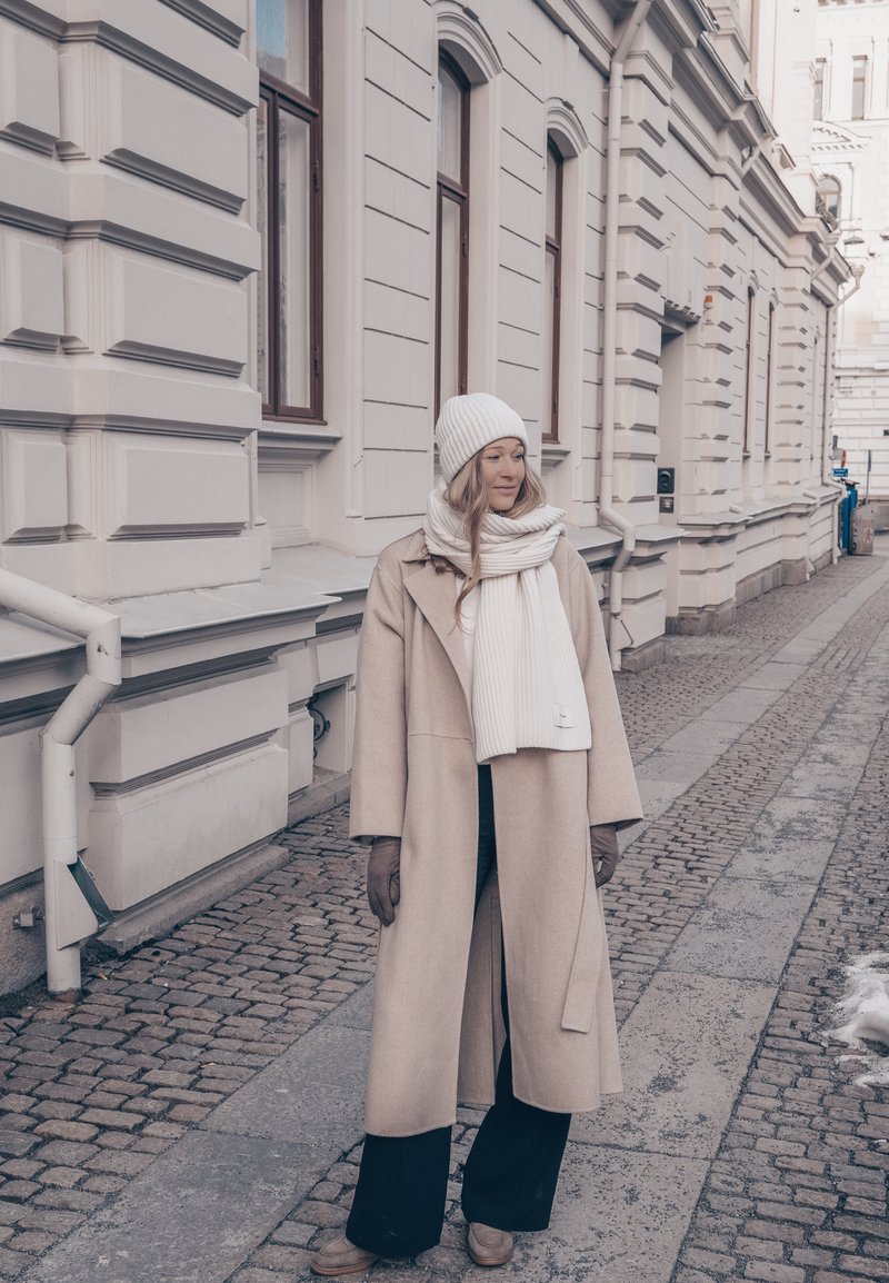 Femme en manteau beige, écharpe blanche et chapeau, debout dans une rue pavée à côté d'un bâtiment clair avec de grandes fenêtres dans un décor hivernal.
