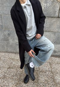 Άτομο με μαύρο παλτό, γκρι πουλόβερ με κουμπιά, σκισμένο ανοιχτό μπλε τζιν, λευκές κάλτσες και μαύρα παπούτσια, που ποζάρει με ένα πόδι σηκωμένο σε πέτρινο πάτωμα.