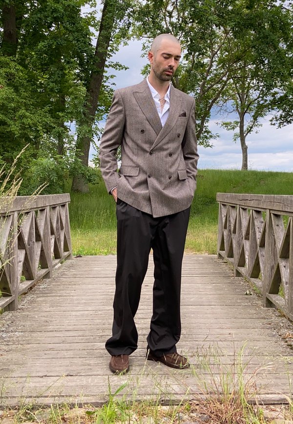 Hombre de pelo corto está de pie en un puente de madera, vistiendo una chaqueta marrón, camisa blanca, pantalones negros y zapatos marrones, rodeado de árboles.