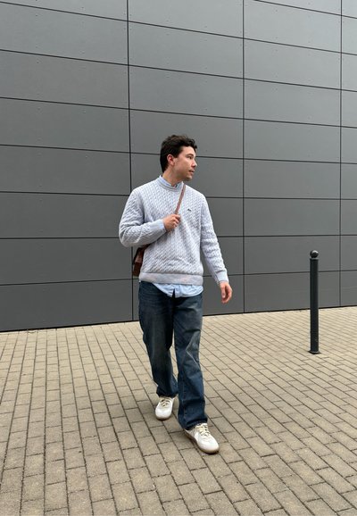 Hombre caminando por una acera con un patrón, vestido de manera casual con un suéter de punto en color gris claro, jeans azules, zapatillas blancas y un bolso de hombro marrón.