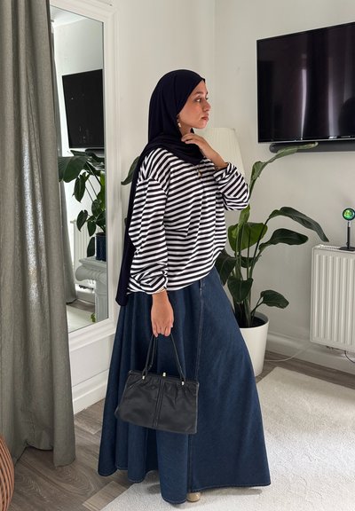 Camisa de rayas de manga larga en blanco y negro, combinada con una falda larga de mezclilla oscura. Sosteniendo un pequeño bolso negro. Entorno interior con vegetación.