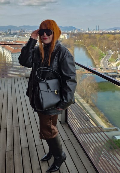 Mujer con cabello rojo que lleva gafas de sol negras de gran tamaño, chaqueta de cuero y botas, está de pie en un balcón con vistas al río y el paisaje urbano.