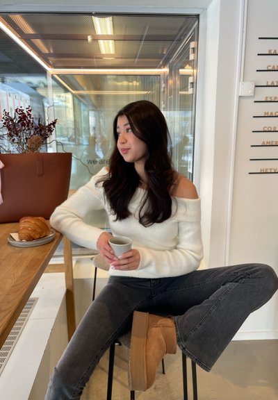 Una mujer vestida con un suéter blanco de hombros descubiertos está sentada en un taburete, sosteniendo una taza gris. A su lado hay una bolsa marrón con un croissant en un plato.