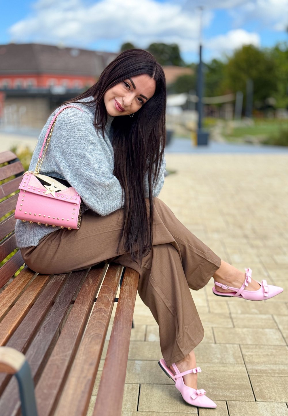 Grå strikket sweater, brune brede bukser, lyserød studset håndtaske og lyserøde slingback sko med sløjfer. Siddende på en træbænk udendørs.