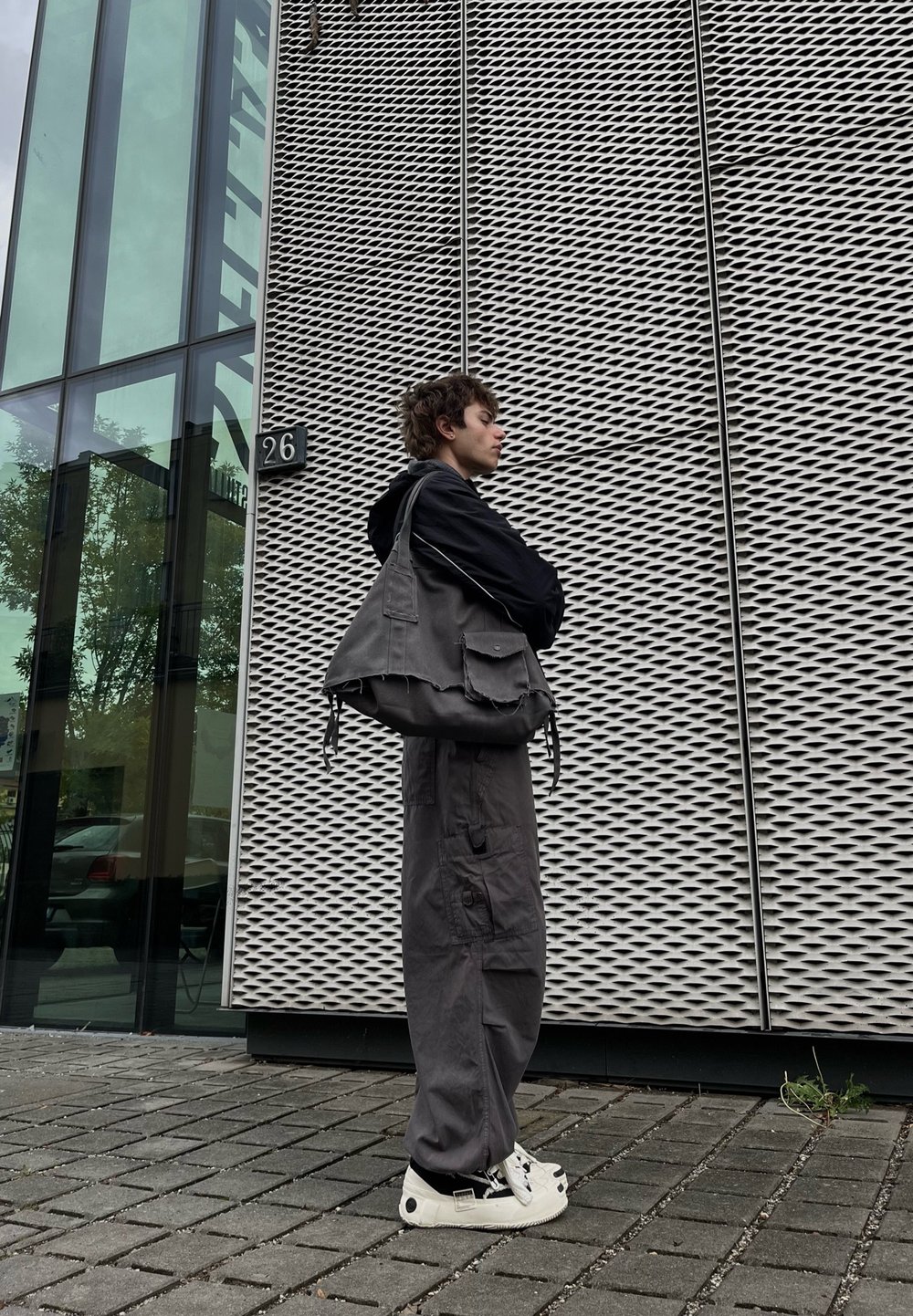 Une jeune personne en vêtements décontractés sombres et avec des baskets blanches se tient contre un mur à motif métallique à l'extérieur d'un bâtiment numéroté 26.