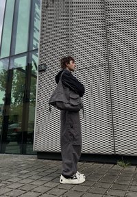 Une jeune personne en vêtements décontractés sombres et avec des baskets blanches se tient contre un mur à motif métallique à l'extérieur d'un bâtiment numéroté 26.