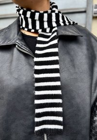 Écharpe tricotée rayée noire et blanche avec une texture douce, présentant un motif côtelé irrégulier, portée sous une veste en cuir noire.
