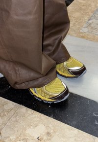 Yellow and black mesh sneakers partially covered by wide brown pants on a polished black and beige tiled floor.