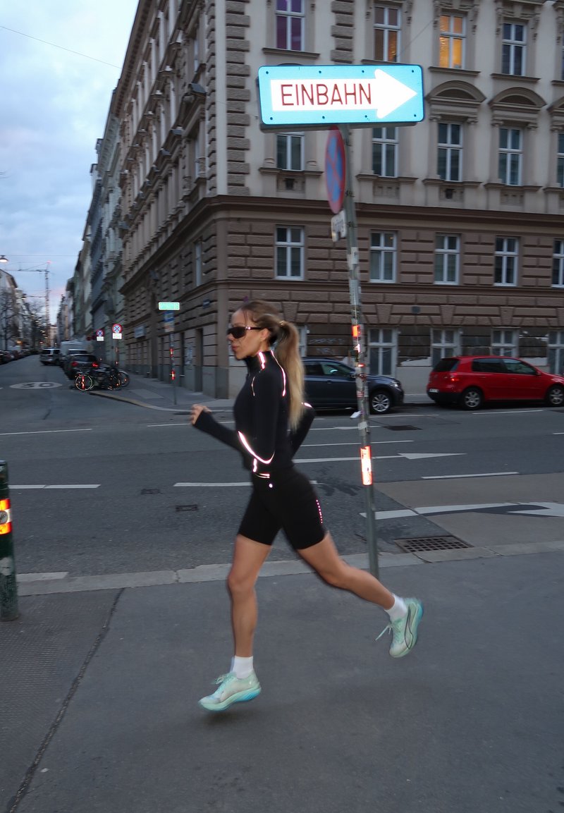 Femme courant en tenue de sport noire avec des bandes réfléchissantes traverse une rue urbaine sous un panneau de sens unique indiquant "EINBAHN."