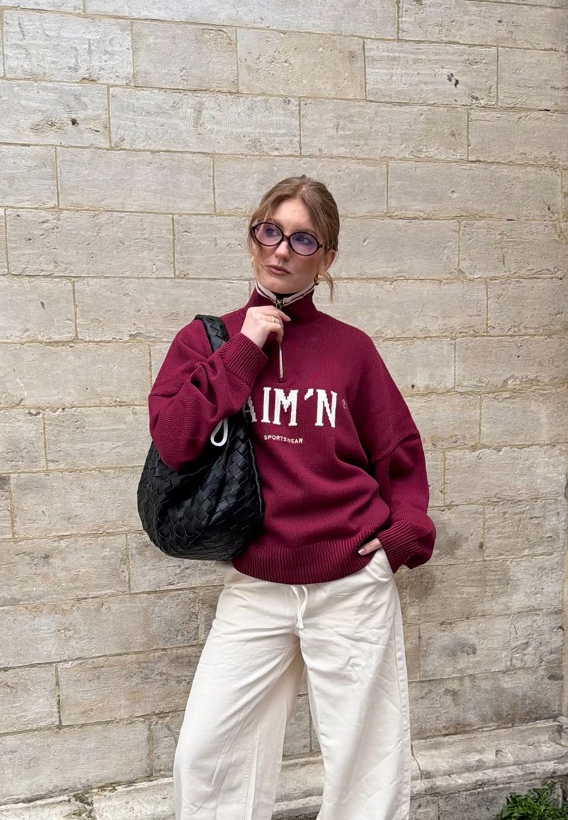 Vrouw met bril, gekleed in een bordeauxrode trui met rits en witte broek, draagt een zwart gevlochten tas en staat tegen een beige bakstenen muur.