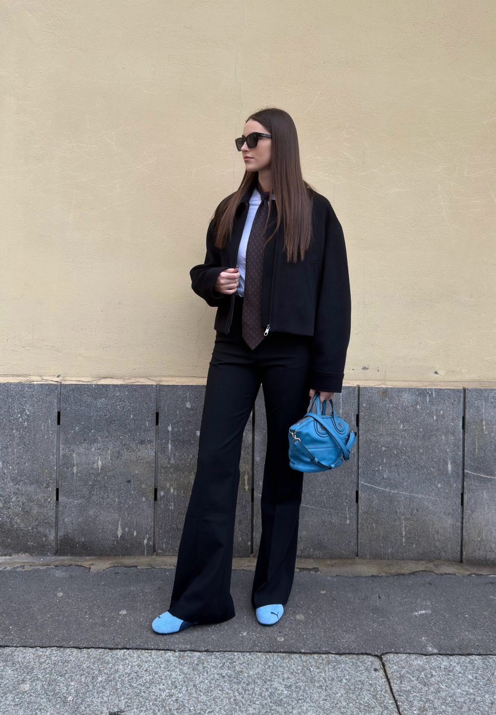 Femme portant une veste noire, une chemise blanche, une cravate brune, un pantalon noir, des chaussures bleues, tenant un sac à main bleu, debout contre un mur beige.
