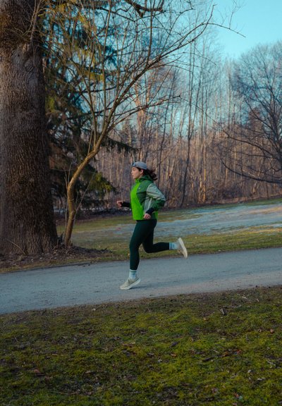 Persona con chaqueta verde y gorra corriendo por un camino de grava en un bosque sin hojas durante el día.