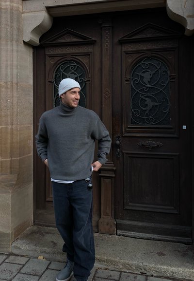 Jersey de cuello alto gris de punto, vaqueros oscuros y holgados, y botas grises. De pie junto a puertas de madera con intrincados acentos metálicos y detalles en relieve.