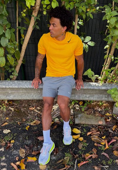 Hombre con camisa amarilla y pantalones cortos grises sentado en una barandilla metálica, rodeado de follaje verde y hojas de otoño caídas en el suelo.