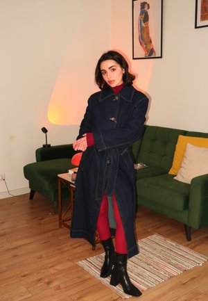 Denim-Trenchcoat mit großem Kragen und Knopfverschluss, kombiniert mit roten Strumpfhosen und schwarzen Absatzstiefeln. Grünes Sofa und gemusterter Teppich im Hintergrund.