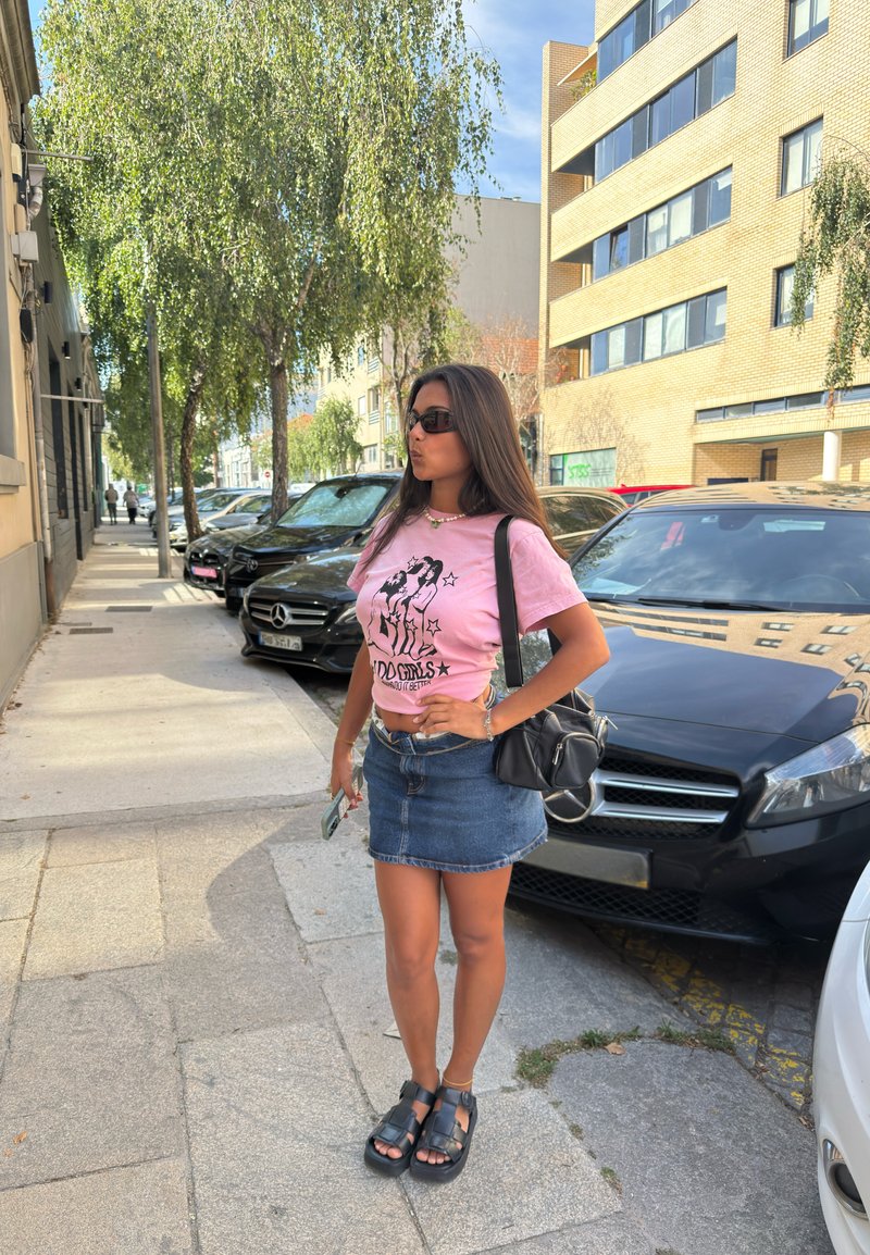 Femme portant des lunettes de soleil, un t-shirt rose avec un motif graphique et une jupe en denim, debout sur un trottoir en ville près de voitures garées et d'arbres.