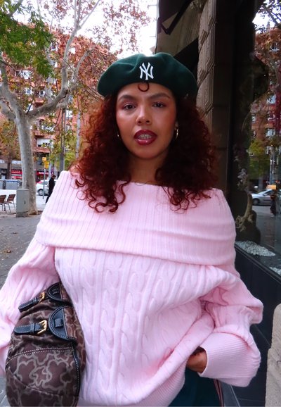 Mujer con pelo rizado que lleva una boina verde de los New York Yankees y un suéter rosa con los hombros descubiertos, parada en una acera de la ciudad cerca de árboles otoñales.