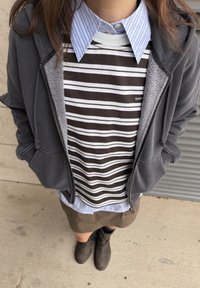 Sudadera gris con cremallera sobre un suéter marrón y blanco a rayas, camisa de cuello azul a rayas, falda beige y botines negros.