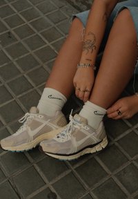 Sapatilhas Nike em tom claro de areia e malha branca com detalhes azuis, combinadas com meias brancas com um logotipo preto, colocadas sobre um piso cinza texturizado.