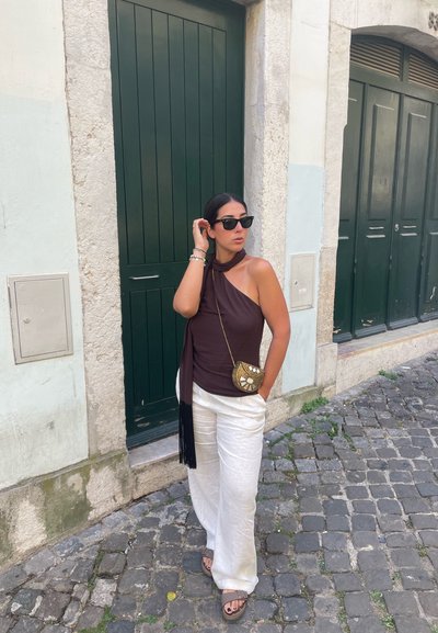 Top sin mangas marrón con detalle de bufanda, pantalones de pierna ancha blancos y un bolso cruzado con detalles dorados, todo contra una calle bordeada de vegetación.