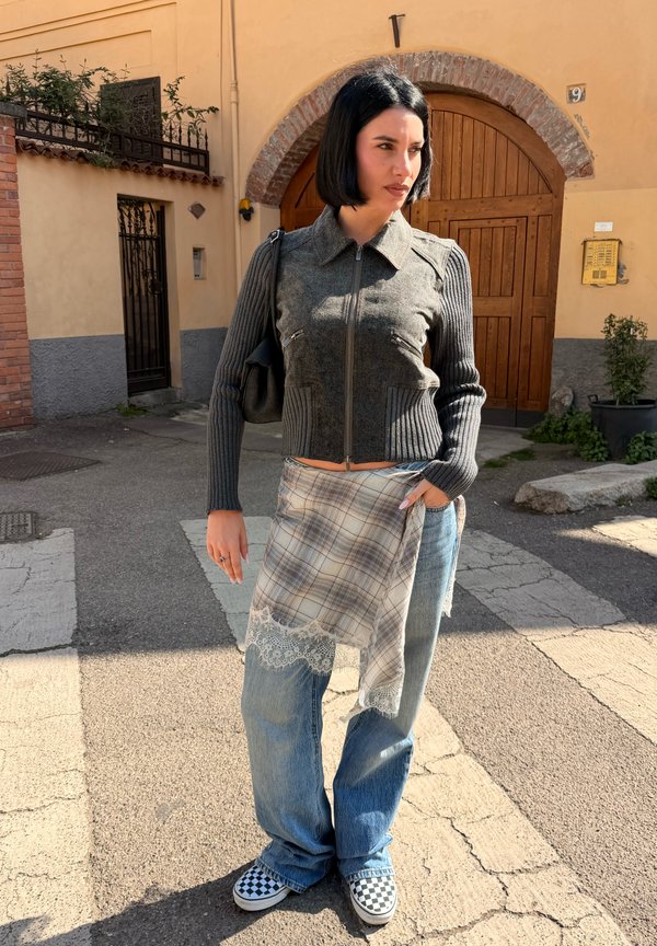 Femme aux cheveux courts noirs debout à l'extérieur portant un pull zippé gris, un tissu à carreaux sur un jean bleu et des chaussures slip-on à carreaux.