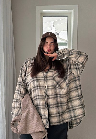 Joven mujer con camisa de cuadros y gorra haciendo una expresión juguetona, sosteniendo una bolsa beige texturizada en el interior cerca de una ventana pequeña.