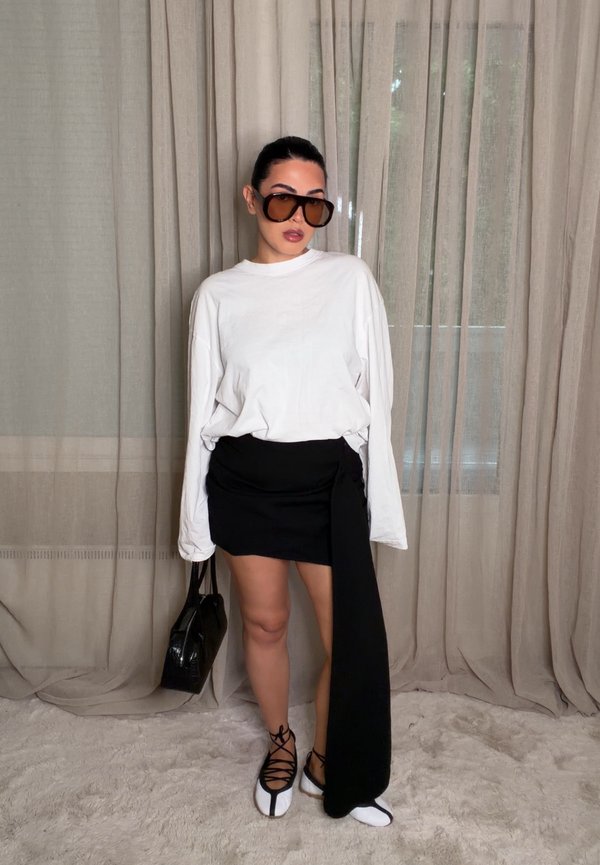 Femme aux cheveux foncés portant un haut blanc à manches longues, une jupe asymétrique noire, des chaussures blanches, de grandes lunettes de soleil et tenant un sac à main noir, debout à l'intérieur.