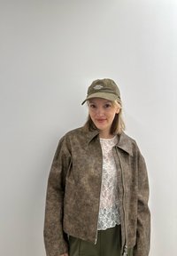 Καφέ ψεύτικο leather μπουφάν με γιακά και κλείσιμο με φερμουάρ, φορεμένο πάνω από ένα λευκό τοπ από δαντέλα, σε συνδυασμό με ένα ελαιοπράσινο καπέλο.