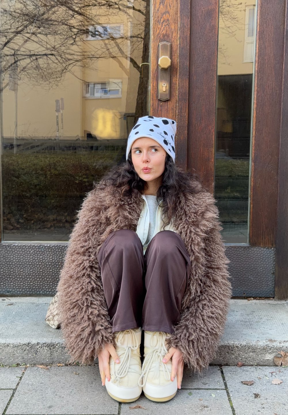 Manteau brun à poils longs, bonnet gris à taches noires, baskets beiges épaisses et pantalon marron. Assis devant une porte en bois.