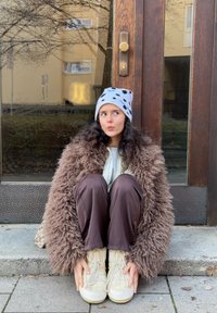 Manteau brun à poils longs, bonnet gris à taches noires, baskets beiges épaisses et pantalon marron. Assis devant une porte en bois.