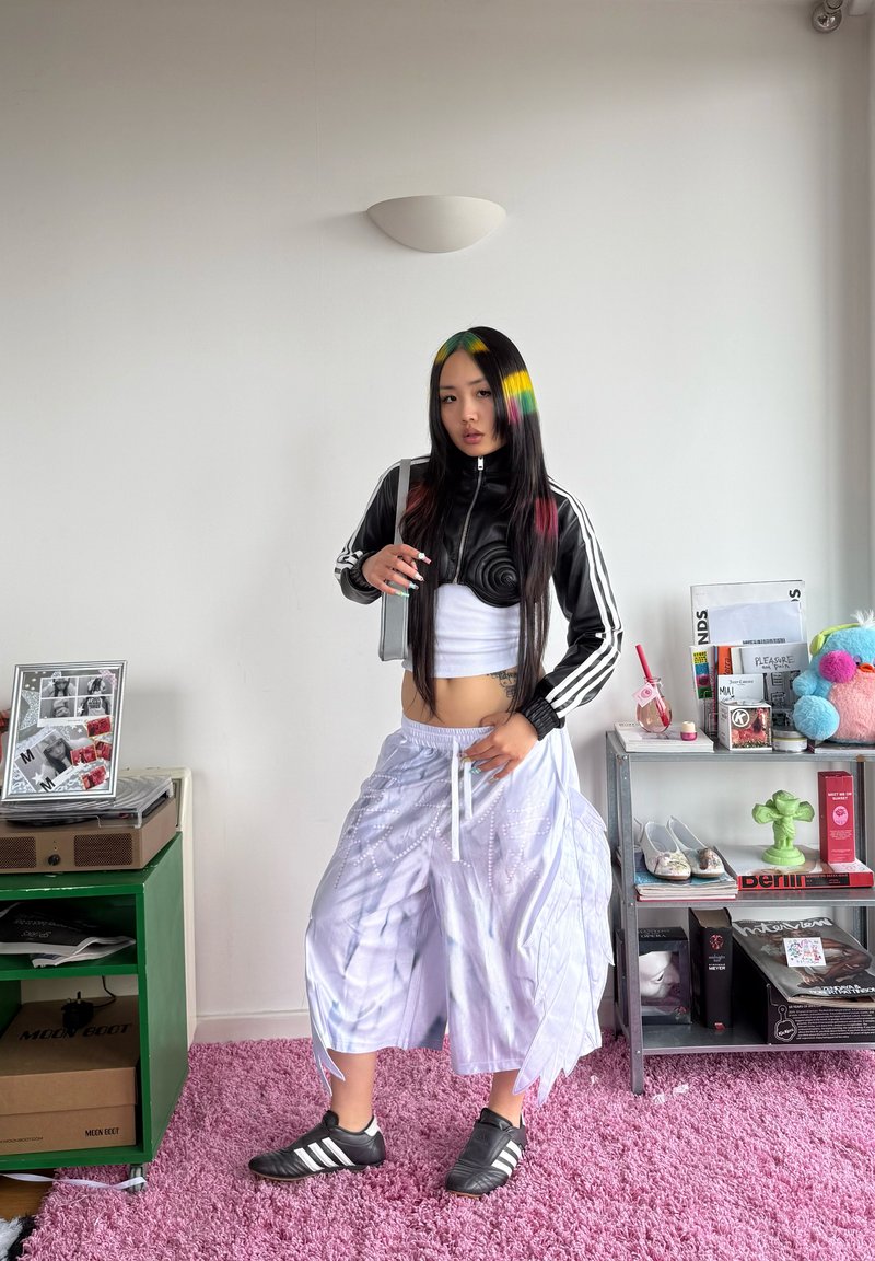 Jeune personne aux longs cheveux noirs teintés de mèches arc-en-ciel, vêtue d'une tenue de sport noire et blanche, debout dans une pièce avec des étagères et un tapis rose.