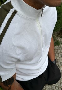 Weißes gestricktes Kurzarm-Polo mit Reißverschlusskragen, ausgestattet mit olivgrünen Seitenstreifen und dezentem Branding-Detail. Strukturierter Stoff mit kurzen Ärmeln.