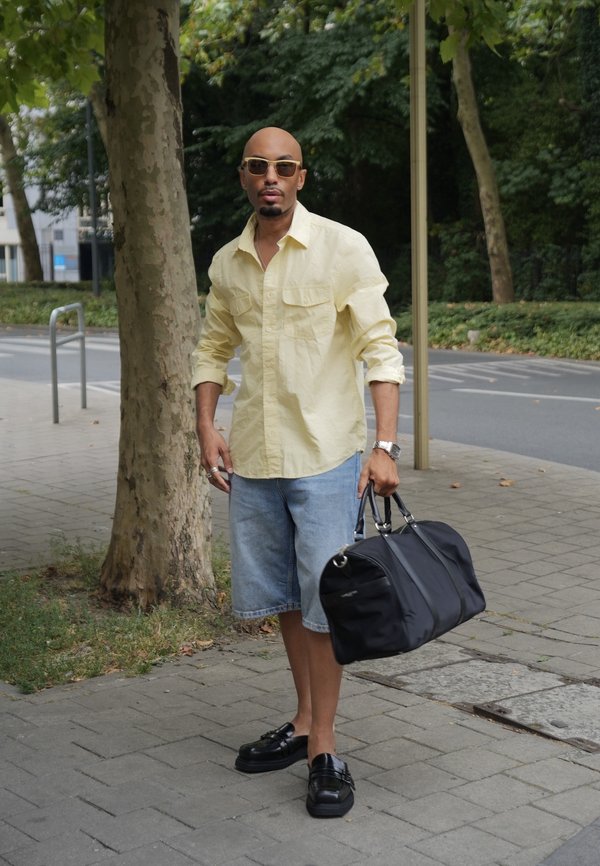 Mand iført solbriller, gul skjorte, denimshorts og sorte sko, der holder en sort sportstaske og står på en fortov nær træer.
