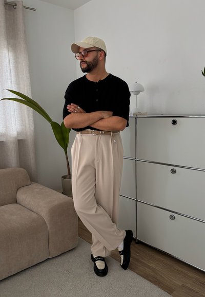 Camisa negra de botones, pantalones beige, zapatos en negro y crema. Diseño minimalista con un cinturón plateado. Entorno interior con tonos claros.
