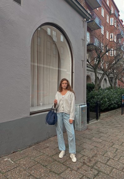 Cárdigan gris claro con botones, top blanco, jeans azul claro y zapatillas blancas. Llevando un bolso azul marino. Fondo urbano con una ventana.