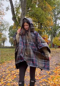 Poncho a quadri in nero, rosso e bianco. Materiale impermeabile con cappuccio. Design largo, indossato sopra una maglia beige e leggings neri, si trova su un sentiero coperto di foglie.