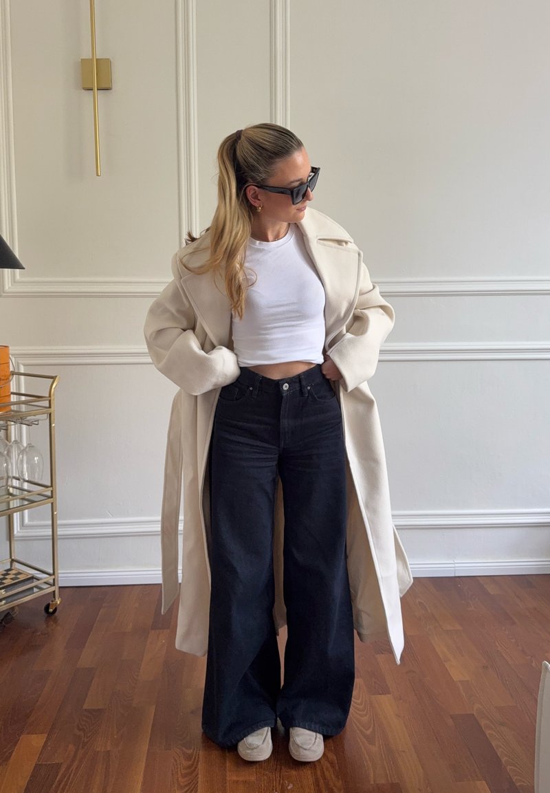 Napszemüveget viselő nő hosszú bézs kabátban, fehér rövid felsőben, bő szárú fekete farmerral és fehér cipőben, beltéren, fapadlón áll.
