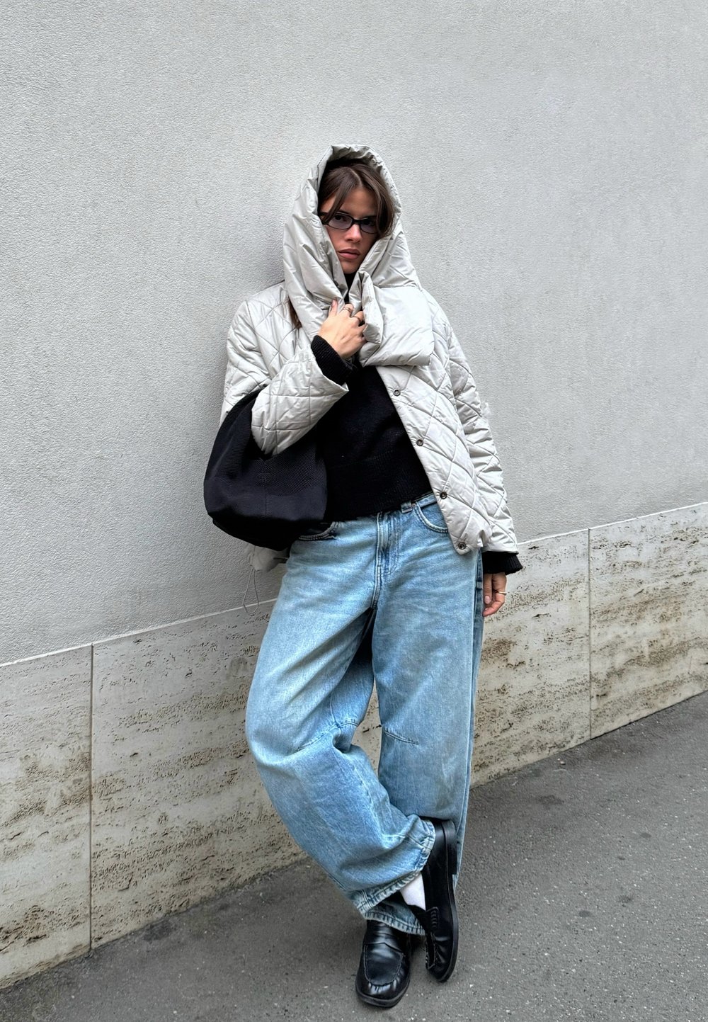 Veste matelassée argentée avec capuche, pull noir et jean bleu clair surdimensionné. Sac noir et mocassins noirs complètent le look.