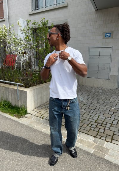 Hombre con gafas de sol ajusta un collar al aire libre cerca de un edificio, lleva camiseta blanca, vaqueros azules, reloj y llaves sujetas al lazo del cinturón.