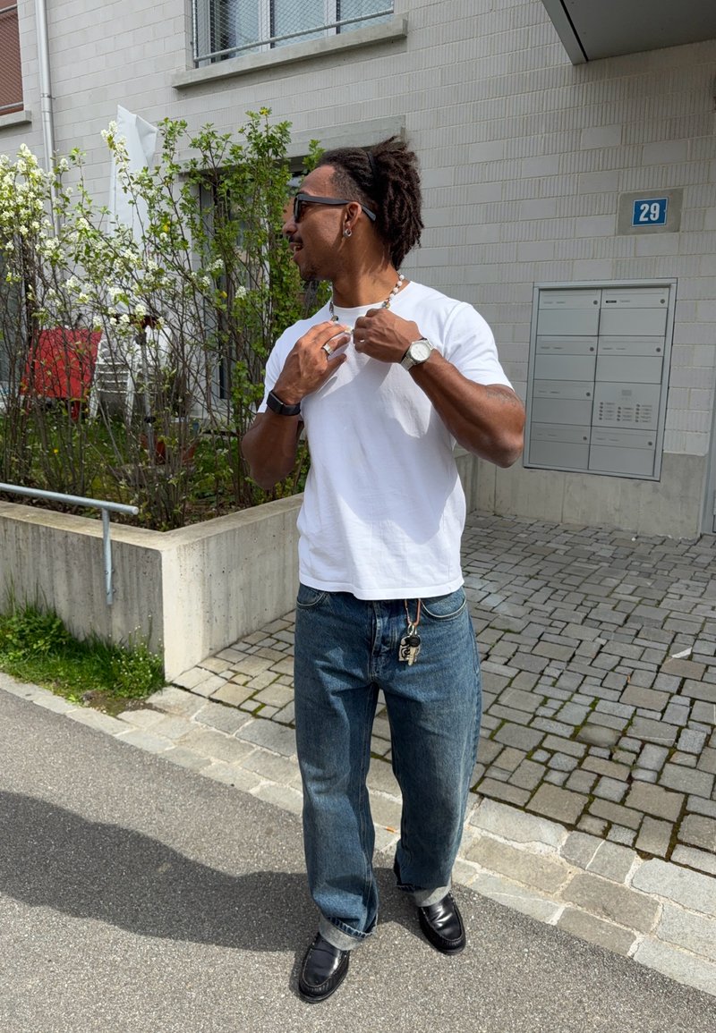 Man met zonnebril past een ketting aan buiten bij een gebouw, draagt wit t-shirt, blauwe jeans, horloge en sleutels vastgemaakt aan de lusselus van de broek.