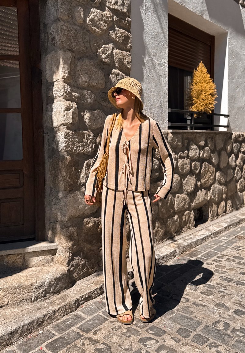 Femme portant une tenue beige et noire rayée, chapeau de soleil et lunettes de soleil, debout dans une rue pavée près d'un mur en pierre avec une petite fenêtre.