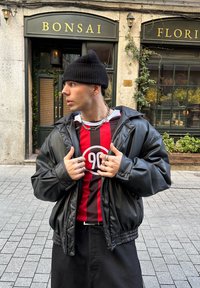 Veste en cuir noire portée sur une chemise rayée rouge et noire avec un motif circulaire. Porte un bonnet en tricot noir et plusieurs bagues, debout en plein air.