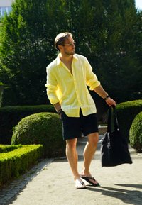 Kollane linane nööbiga särk, tumesinised lühikesed püksid, mustad plätud, must kott käes. Taustal looduslik rohelus.