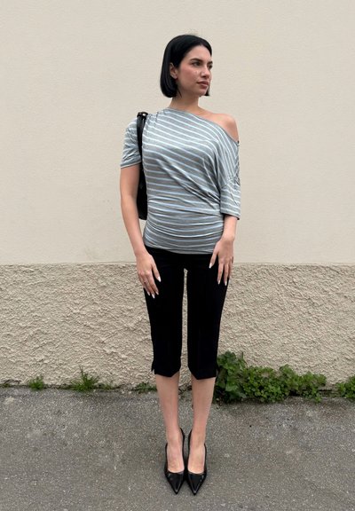 Mujer con pelo corto y negro de pie contra una pared beige, lleva una blusa de rayas sin hombros, pantalones negros cortos, tacones negros y lleva un bolso negro.