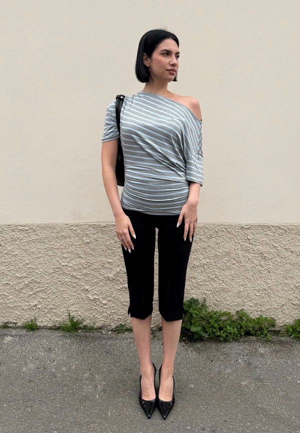 Sieviete ar īsiem melniem matiem stāv pie bēša krāsas sienas, valkājot svītru off-plecu topu, melnas īsas bikses, melnas kurpes ar papēžiem un nēsājot melnu somu.